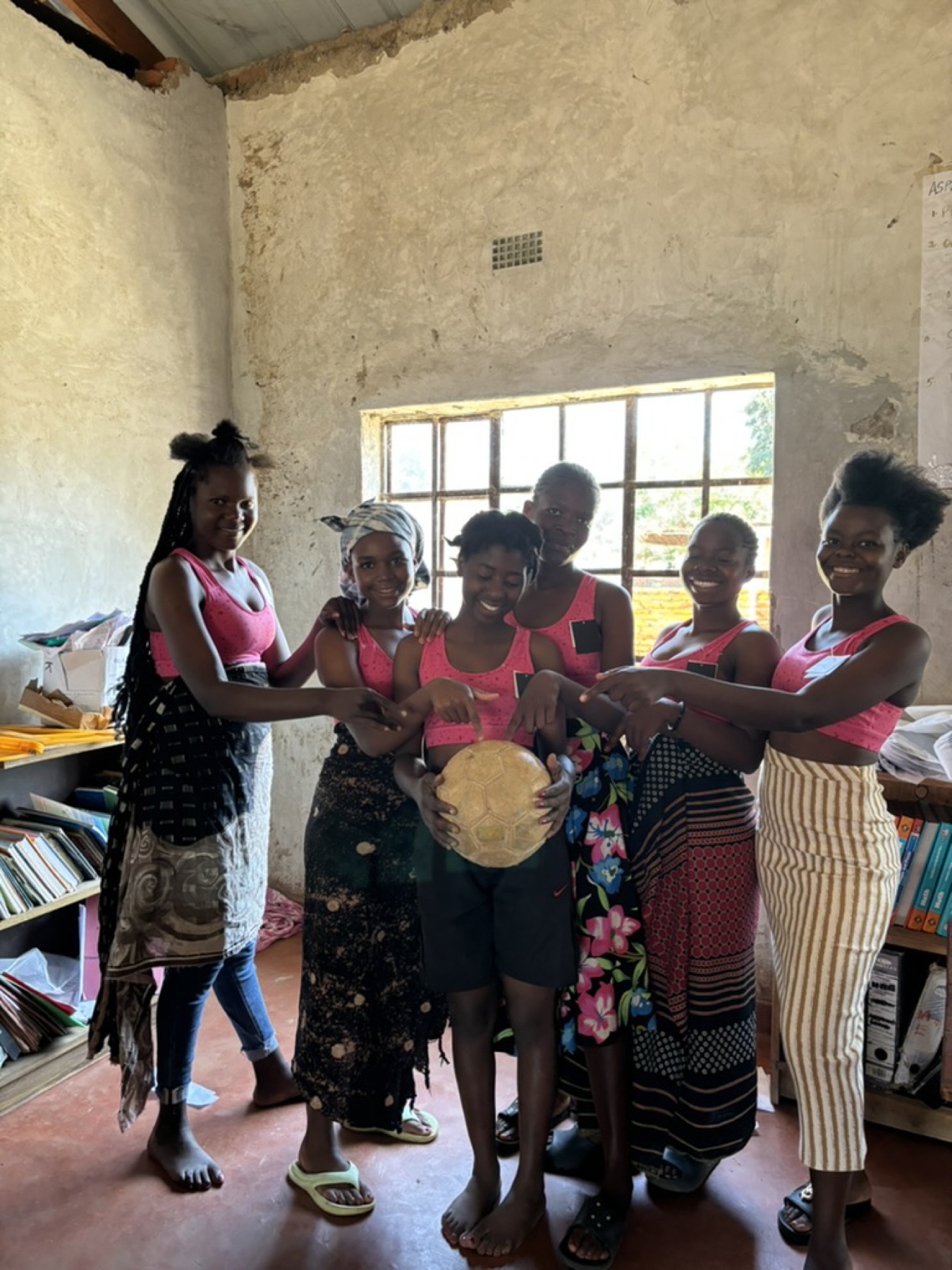 Six girls standing together indoors, hands on a football they are holding between them