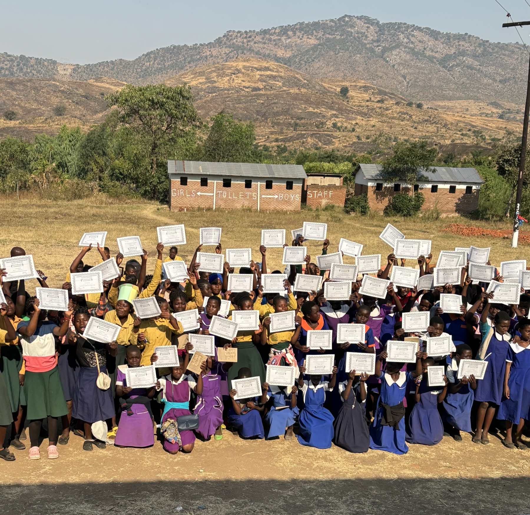 A whole school holds up I Am Who? certificates outside, with the hills behind them