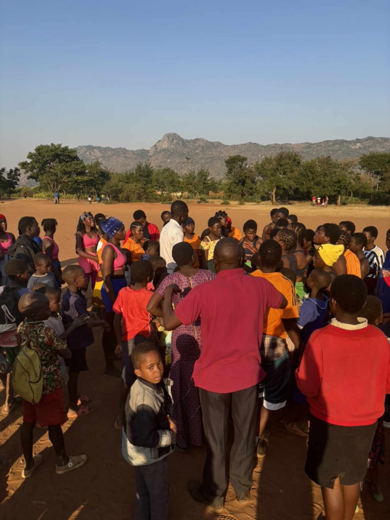 A crowd of children and adults gathered on open ground at dusk, hills behind them